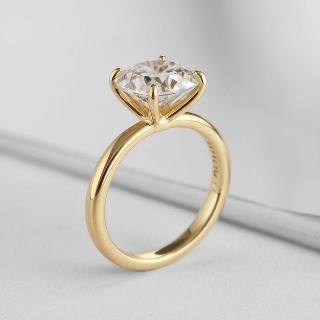 A close-up of a classic solitaire gold ring featuring a brilliant diamond, set against a soft, neutral background. The simplicity of the design highlights the stone's sparkle.