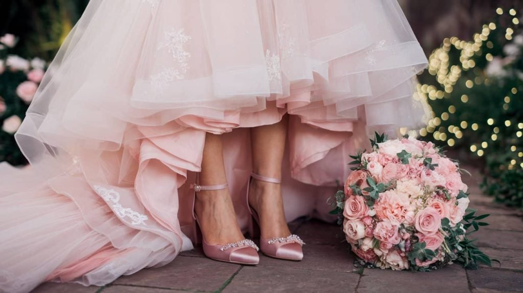 A delightful image showcasing a bride in a breathtaking blush wedding dress with intricate lace details and a flowing tulle skirt. The focus is on her elegant blush pink heels, adorned with delicate embellishments, paired with a stunning bouquet of soft pink roses, peonies, and greenery. The gown drapes gracefully around her as she stands in a romantic garden setting, with sunlight filtering through lush foliage, adding to the enchanting ambiance.