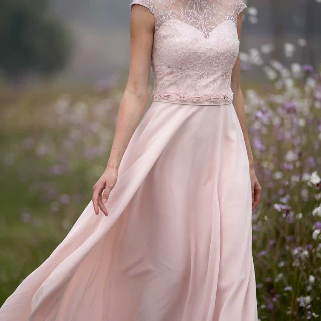 Bride in a pastel pink blush wedding dress, walking through a field of wildflowers with her gown swaying in the breeze.