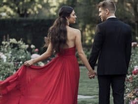 A beautiful woman in a red flowy Valentine's Day dress with a half-up, half-down hairstyle, holding hands with her handsome date in a black suit, set against a lush garden backdrop filled with blooming flowers.