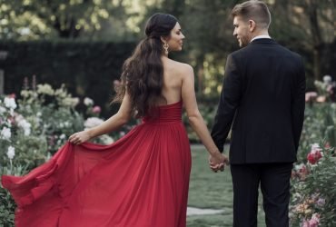 A beautiful woman in a red flowy Valentine's Day dress with a half-up, half-down hairstyle, holding hands with her handsome date in a black suit, set against a lush garden backdrop filled with blooming flowers.