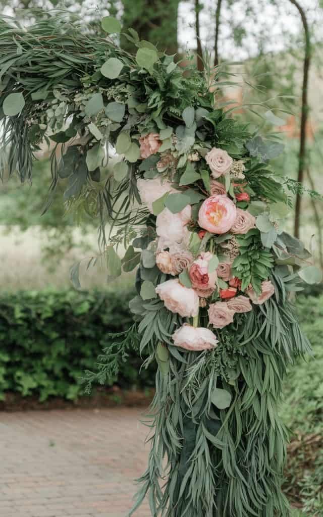 Charming Wedding Arch Flowers incorporating a mix of greenery and elegant flowers like garden roses, adding depth and texture to an outdoor wedding setting.
