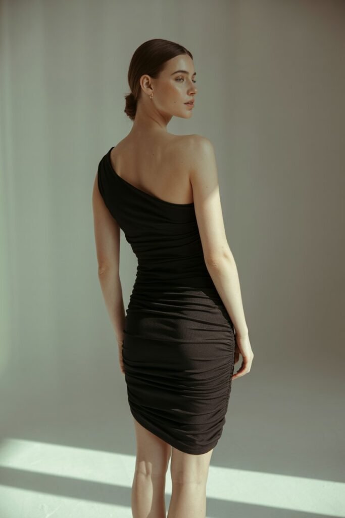 A full-length editorial photo of a woman with her back slightly turned, wearing a one-shoulder black ruched jersey cocktail dress. She is in a minimalist, brightly lit studio with a soft grey background. The lighting highlights the draping of the fabric. Portrait aspect ratio (2:3)