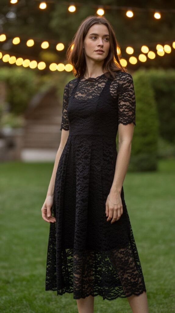 A woman is standing in a softly lit garden at dusk, fairy lights blurred in the background. She is wearing a black lace A-line midi dress, and the camera captures the delicate texture of the lace. Portrait aspect ratio (2:3).